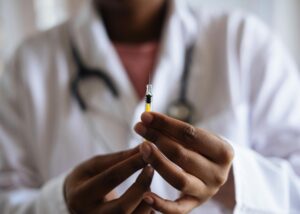 A imagem mosta um enfermeiro negro segurando com duas mãos uma seringa com líquido que se assemelha a vacina da tuberculose